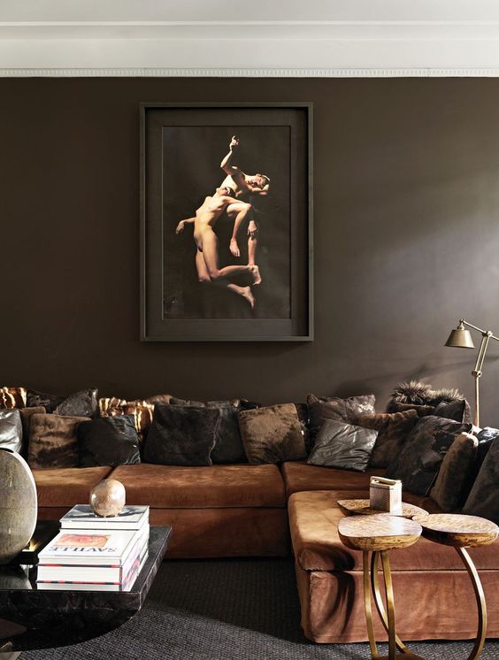 A moody and exquisite liivng room with dark brown walls, a rust colored sofa and pillows, a coffee table and some lamps