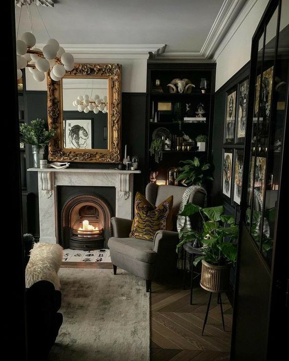 A moody modern Goth living room with a fireplace, a grey chair, built in shelves with decor, a mirror in a gold frame, potted greenery