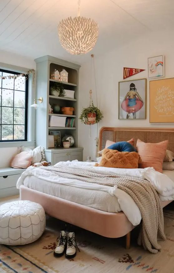 a muted color boho bedroom with a pink bed with a rattan headboard, pale green storage unit with a window seat and a chandelier