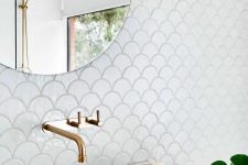 a neutral bathroom clad with white fishscale tiles, a sink clad with stone, gold fixtures and a round mirror is cool