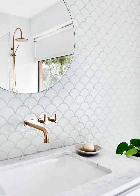 a neutral bathroom clad with white fishscale tiles, a sink clad with stone, gold fixtures and a round mirror is cool