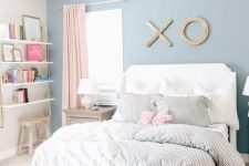 a pastel teen bedroom with a blue accent wall, a white bed with printed bedding, a blue upholstered bench, open shelves, a gold chandelier