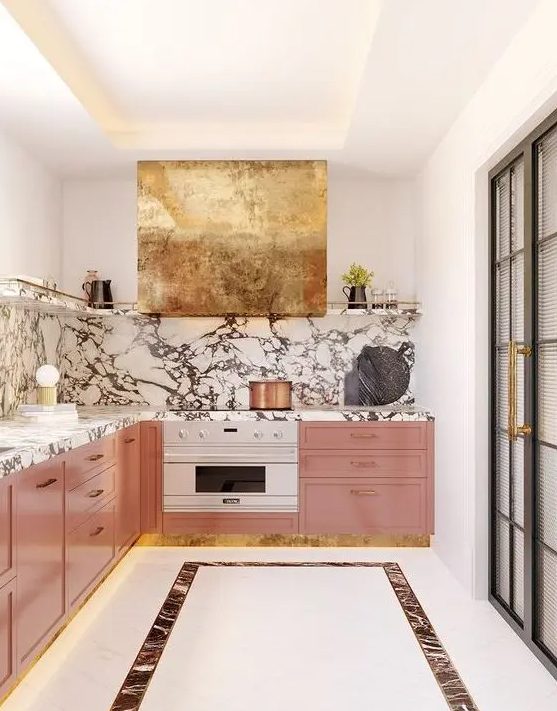 a pink kitchen with a white stone backsplash and countertops plus a shiny gold hood for a super glam accent