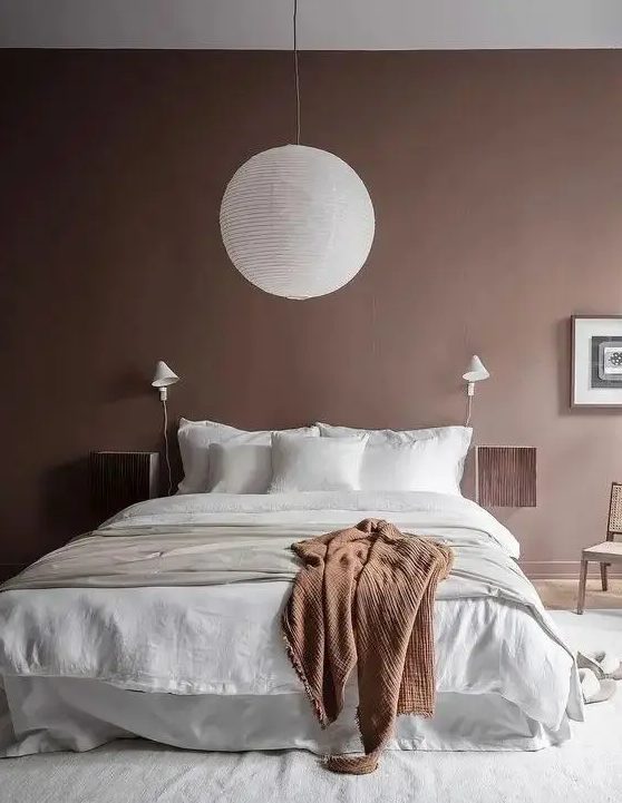 a pretty bedroom with brown walls, a bed with neutral bedding, floating nightstands, a pendant lamp and a chair