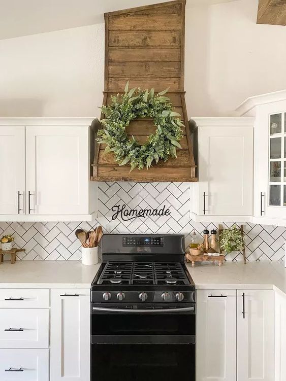 a pretty farmhouse kitchen in white, with a herringbone tile backsplash and a wooden hood with a greenery wreath