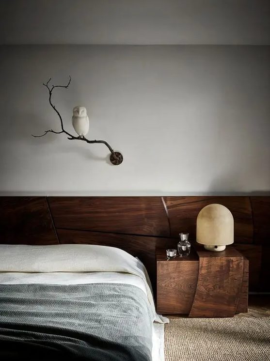 A refined contemporary bedroom clad with dark stained wood, with a low bed, a nightstand, a table lamp and a branch with an owl as decor