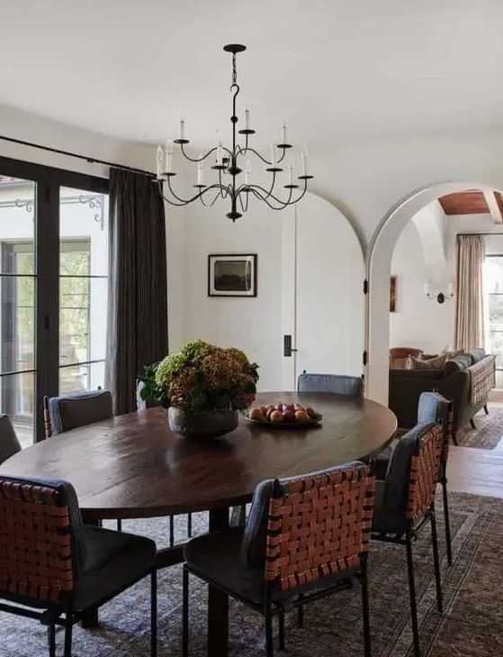 A refined dining room with an oval dark stained table, black woven chairs, potted greenery and a vintage chandelier