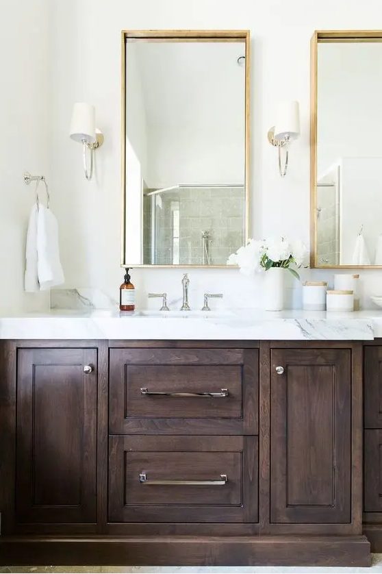 A refined neutral bathroom with a dark stained vanity, a white stone countertop, mirrors in gilded frames and neutral sconces