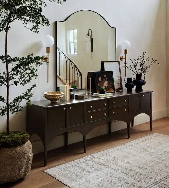 A refined vintage dark stained console table with artwork, books and vases, a large mirror and a potted tree next to it