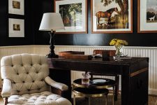 a refined vintage home office with black walls and creamy paneling, a dark-stained desk, a creamy chair and coffee tables