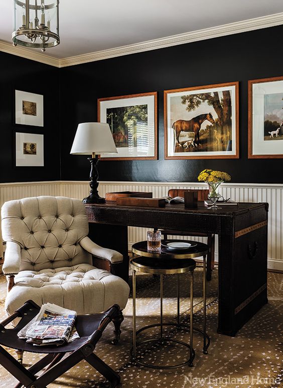 A refined vintage home office with black walls and creamy paneling, a dark stained desk, a creamy chair and coffee tables