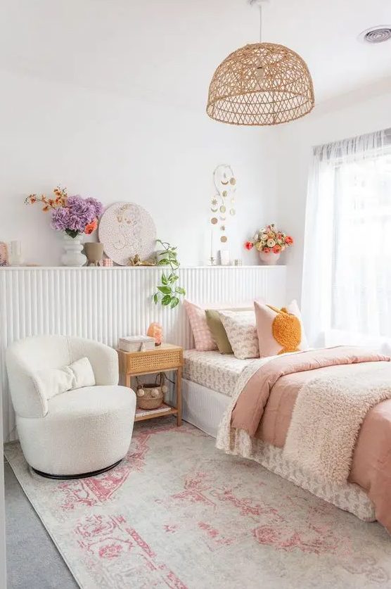 a romantic girl's bedroom with a paneled shelf with decor and blooms, a bed with pastel bedding, a creamy chair and a woven pendant lamp