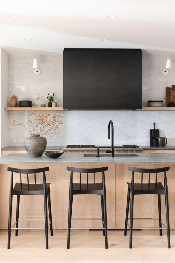 A serene Japandi kitchen with light stained cabinets, grey stone countertops, black stools, fixtures and a black hood