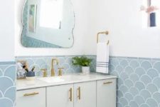 a sink space done with blue fish scale tiles, a vanity, gold fixtures and sconces and a quirky-shaped mirror