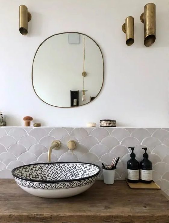 a small and elegant sink space with grey scallop tiles, a printed sink, a mirror and catchy brass tube lamps