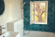 a small bold bathroom done with white and turquoise fish scale tiles, white appliances, a small window and a round mirror