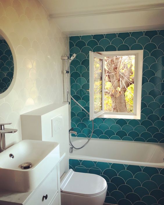 a small bold bathroom done with white and turquoise fish scale tiles, white appliances, a small window and a round mirror