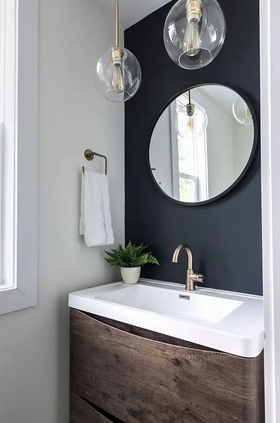a small powder room with a soot accent wall, a timber vanity with a stone sink, a round mirror and pendant lamps
