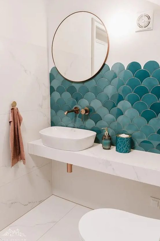 a small sea-inspired bathroom clad with white marble tiles and blue and turquoise fish scale ones, a vanity with a sink and a round mirror