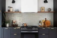 a soot kitchen with gold handles, a white hood, open shelves, black sconces and decor, artwork is a stylish idea