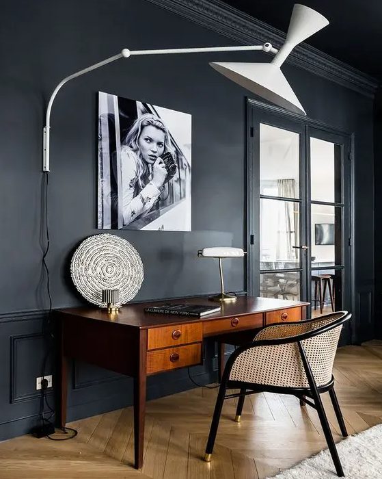 a soot space with a vintage rich stained desk and a cane chair, a large wall lamp and an artwork