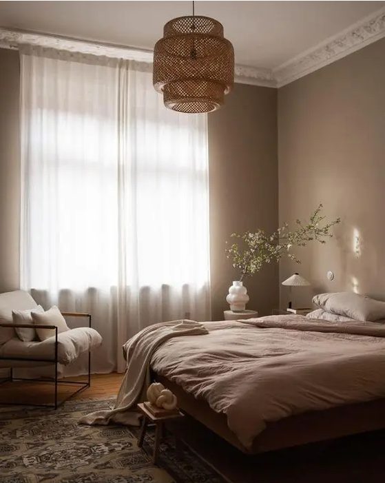 a soothing bedroom with light brown walls, a bed with neutral bedding, a neutral chair and a woven pendant lamp