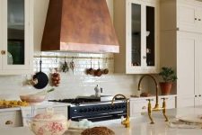 a sophisticated kitchen with creamy cabinets, an olive green kitchen island, an aged metal hood and a white tile backsplash