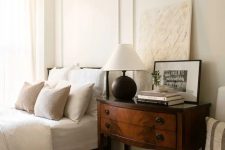 a sophisticated neutral bedroom with creamy bedding, a vintage dark-stained nightstand, some decor and a chair