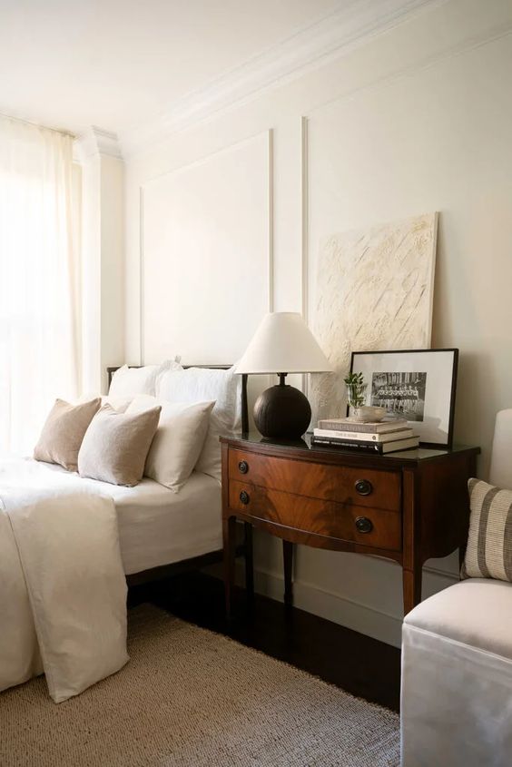 A sophisticated neutral bedroom with creamy bedding, a vintage dark stained nightstand, some decor and a chair