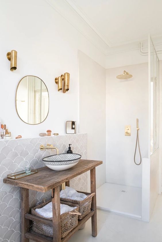 a stylish neutral bathroom with a grey fish scale tile wall, a rough wood vanity, gold fixtures and some baskets