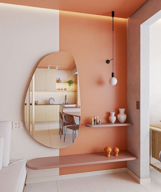 A stylish nook with a Peach Fuzz accent, built in light, some shelves, a creatively shaped mirror and some decor