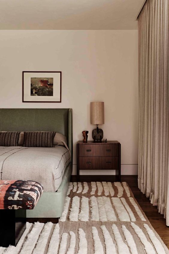 a subtle earthy bedroom with greige drapes and a taupe rug, a green bed with neutral bedding, nightstands and a bold bench