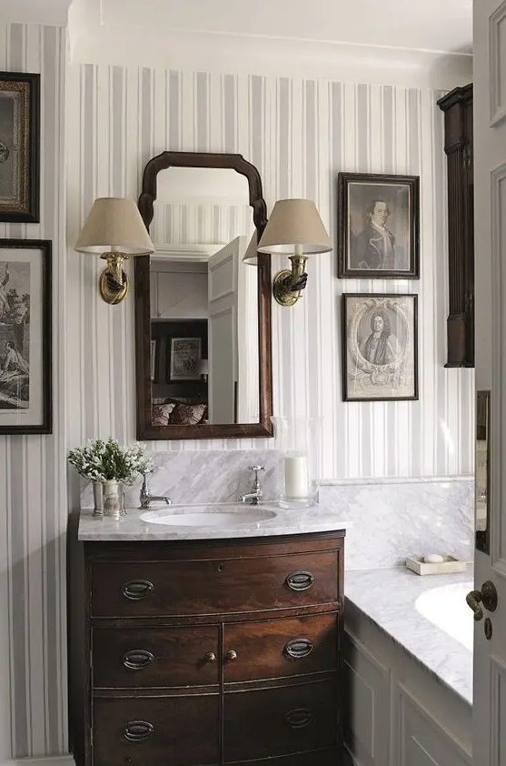 A vintage bathroom done with striped wallpaper, a dark stained vanity, a gallerywall and a mirror in a stained frame, a couple of vintage sconces