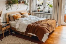 a welcoming earthy boho bedroom with stained furniture, rust and white bedding, a patterned rug, lots of potted plants and a woven pendant lamp