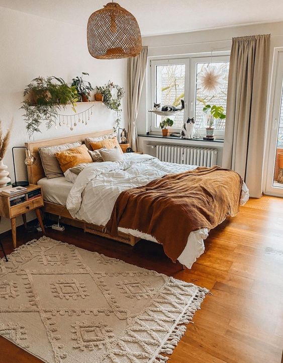 a welcoming earthy boho bedroom with stained furniture, rust and white bedding, a patterned rug, lots of potted plants and a woven pendant lamp
