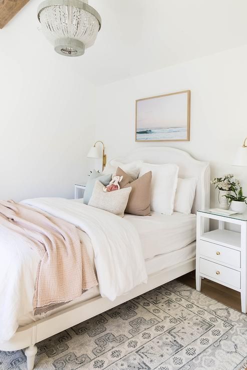 a white French twin bed with pale pink blanket, a white nightstand and a white and gray rug, illuminated by a white and gray beaded chandelier