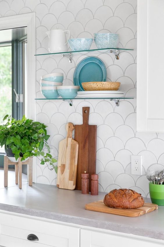 a white kitchen with grey stone countertops, a white scallop tile backsplash, clear glass shelves and greenery