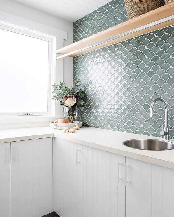 a white planked kitchen with a grey fishscale tile backsplash, an open shelf and some greenery for a seaside feel