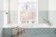 an airy coastal bathroom done with blue fish scale tiles, a tub by the window, a small stool and a lot of natural light