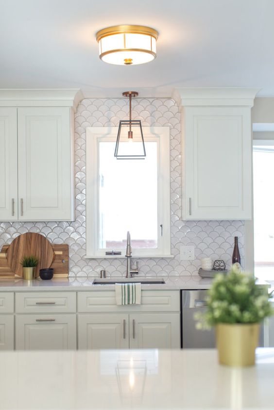 an airy white kitchen with a white fish scale tile backsplash and white stone countertops plus lights and greenery