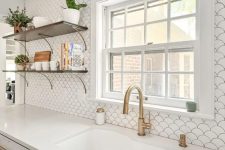 an airy white kitchen with shaker cabinets, a white fishscale tile backsplash, open shelves and brass fixtures and lamps