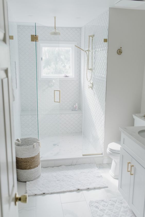 an airy whte bathroom done with white fish scale tile in the shower, a window, a white vanity, white towels and a basket