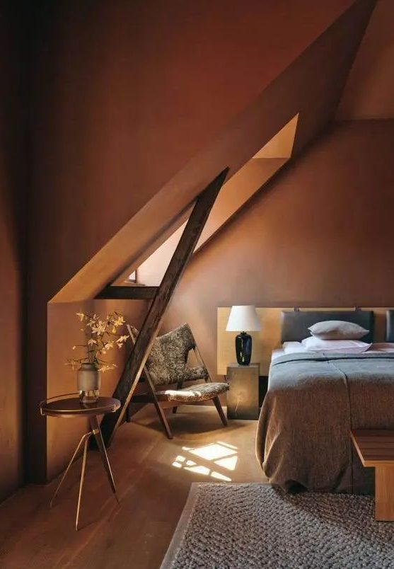 an earthy attic bedroom with burgundy walls, a bed with neutral bedding, stone nightstands and an eye-catchy chair