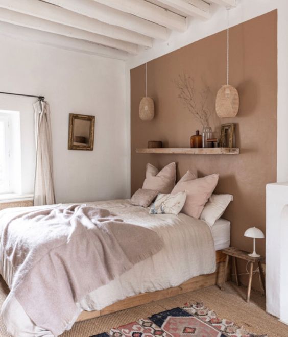 an earthy bedroom with a terracotta wall, a bed with neutral bedding, a shelf with some decor and a bold printed rug