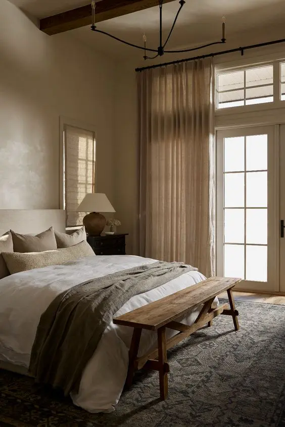 an earthy bedroom with tan walls, a modern neutral bed with neutral bedding, a wooden bench and neutral textiles