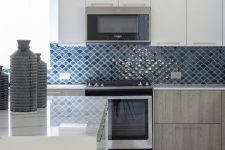 an elegant white and stained kitchen with blue and navy fish scale tile backsplash, white stone countertops and pendant lamps