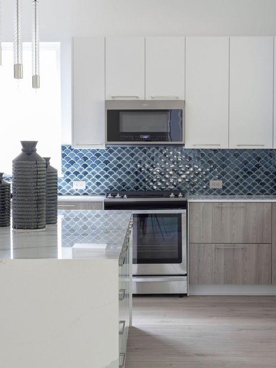 an elegant white and stained kitchen with blue and navy fish scale tile backsplash, white stone countertops and pendant lamps
