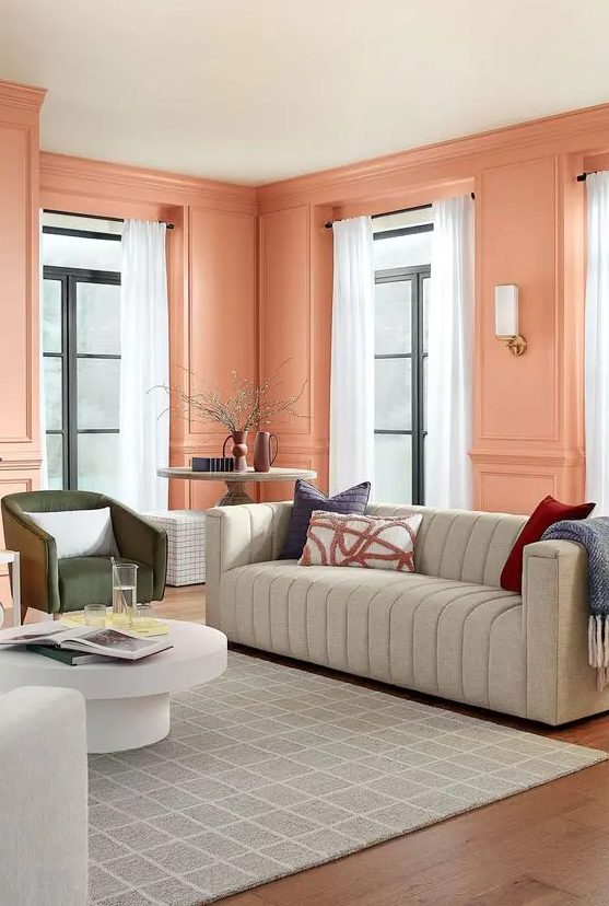 An eye catchy peach pink living room with paneled walls, a grey sofa, a green chair, a coffee table and neutral curtains