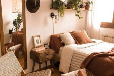 an inviting earthy bedroom with a bed with rust and neutral bedding, a basket with pillows, a woven chair and some potted plants