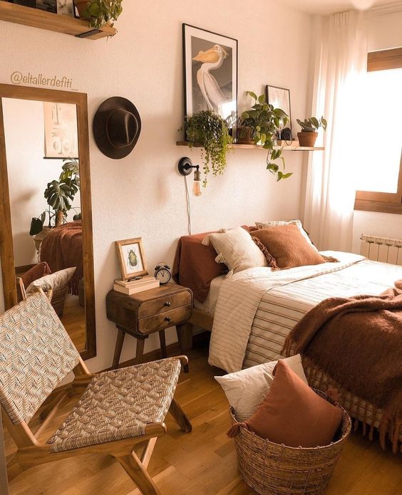 an inviting earthy bedroom with a bed with rust and neutral bedding, a basket with pillows, a woven chair and some potted plants